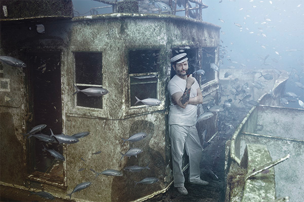 Man smokes pipe on shipwreck surrounded by fish.