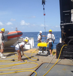 A crew on the deck of a ship pulls a remotely operated vehicle out of the water with a cable.